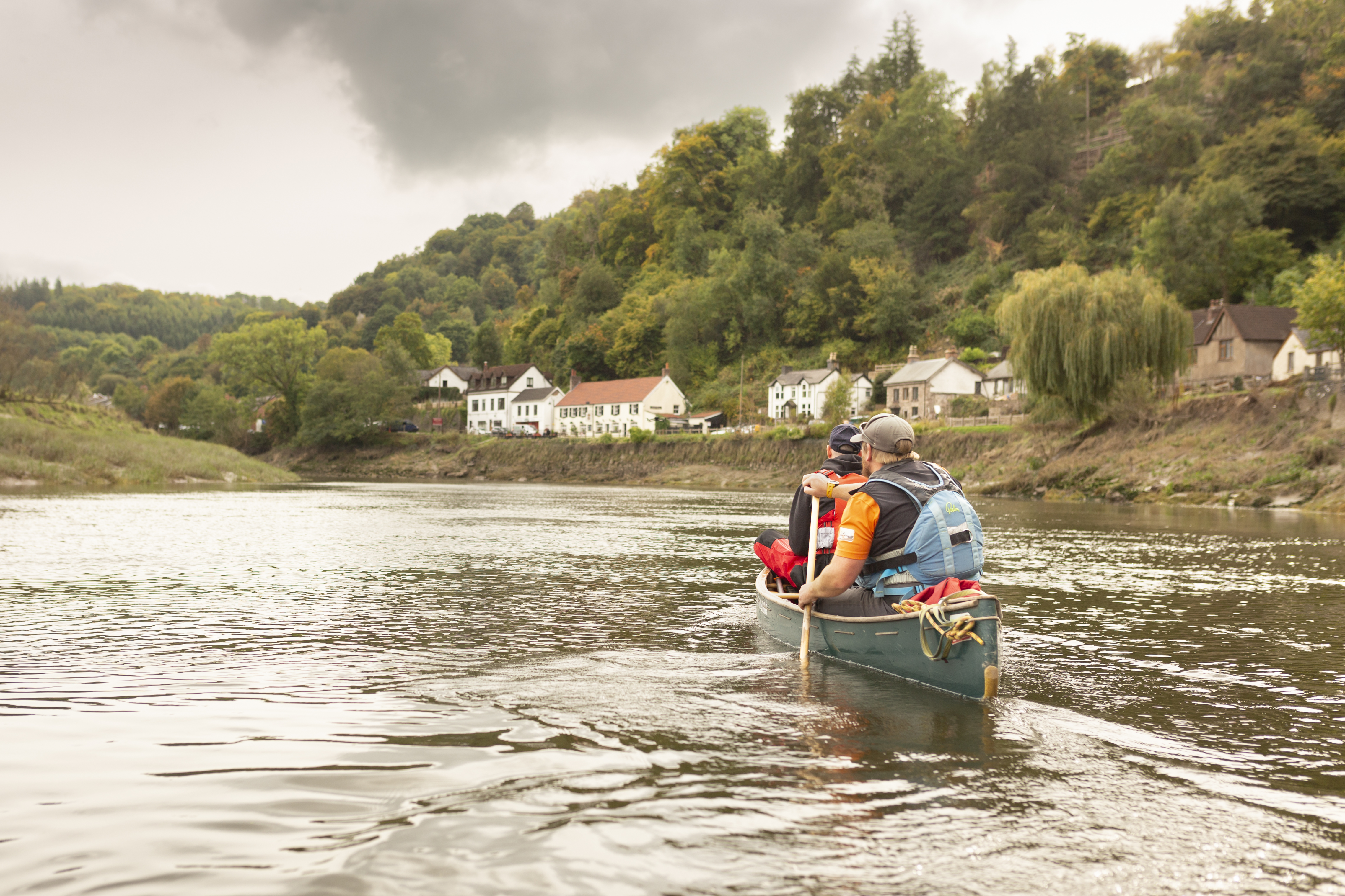 Two people canoe on a calm river with houses and forested hills under a cloudy sky in the background.