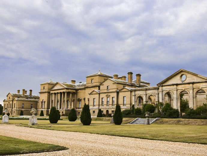 Large neoclassical mansion set in landscaped gardens with a gravel path, manicured lawns, and overcast sky.