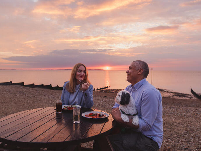 Una donna e un uomo con un cane seduti su una panchina da picnic su una spiaggia al tramonto