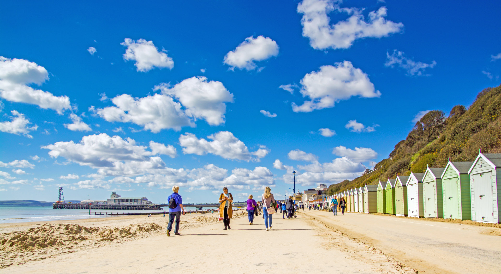 People walking along the coast next to colourful beach huts on a sunny day.
