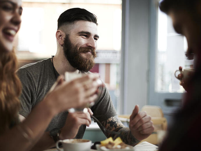 Jeune homme barbu et tatoué discutant avec un groupe d'amis dans un café.