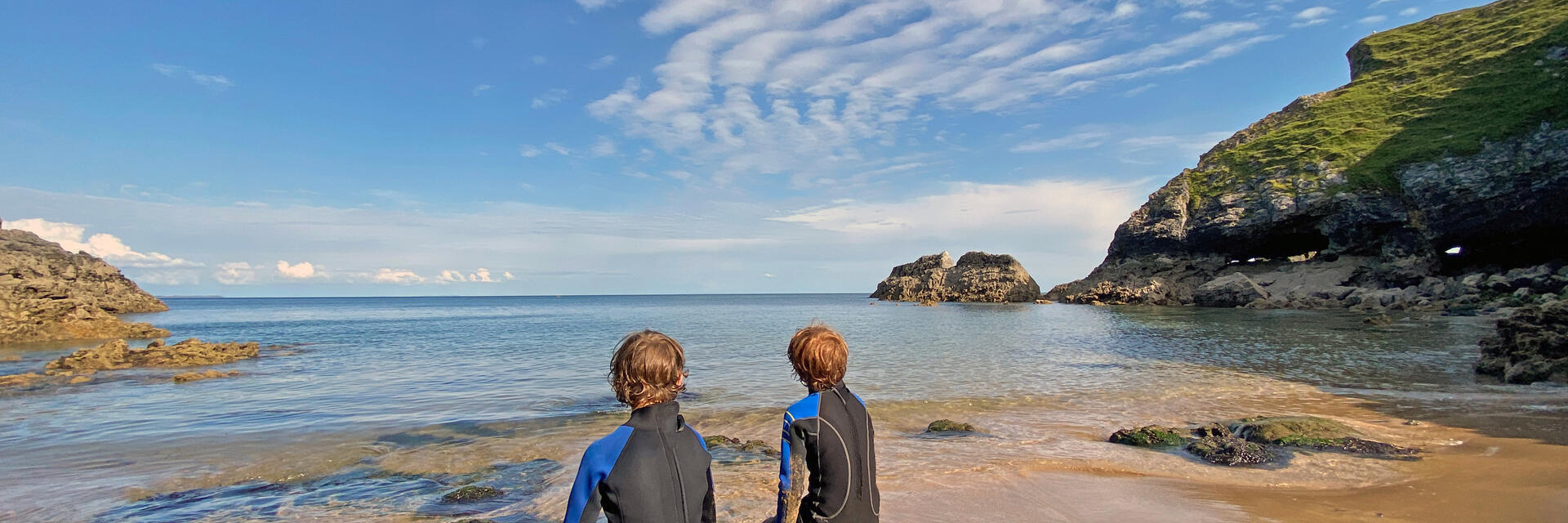 Back view of two young boys in wetsuits sitting on a rock looking out to sea with blue sky