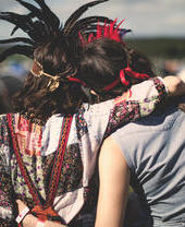 Two people in colorful outfits and feather headbands sit on grass at an outdoor festival, arms around each other, tents in the background.