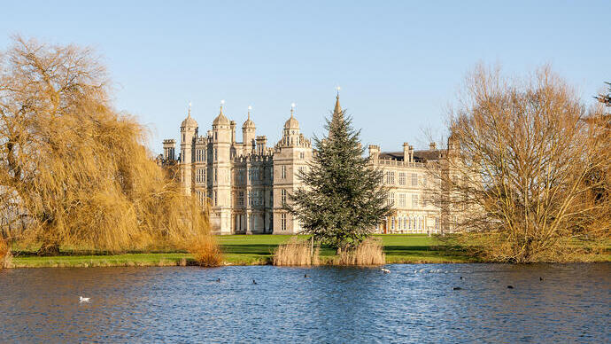 Grand bâtiment historique avec des tours ornées vu à travers un lac, entouré d’arbres et de pelouses vertes sous un ciel bleu clair.