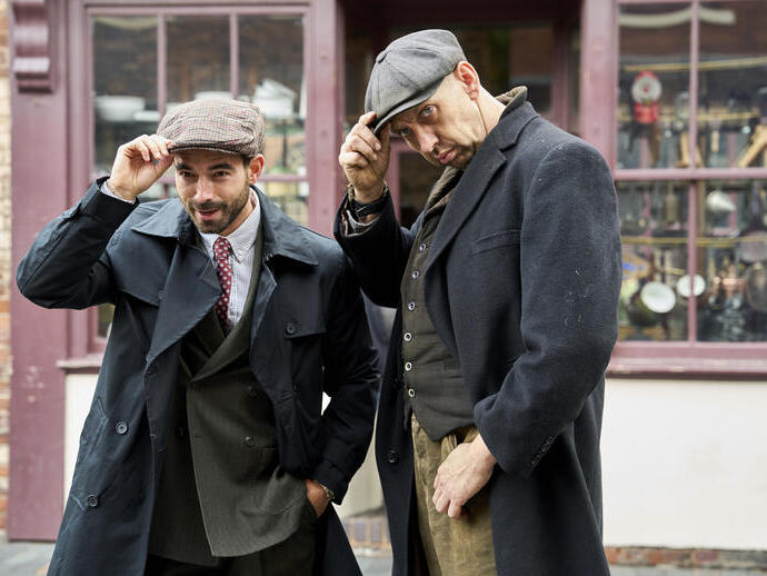 Two men in vintage clothing and flat caps pose outside a shop with a display window and brick wall backdrop.