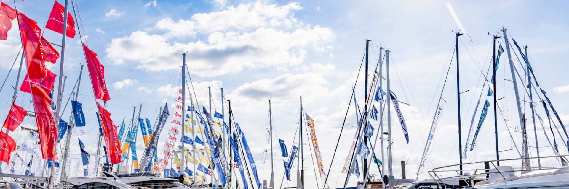 Several sailboats with colorful flags docked at a marina under a bright sky with sun and scattered clouds.