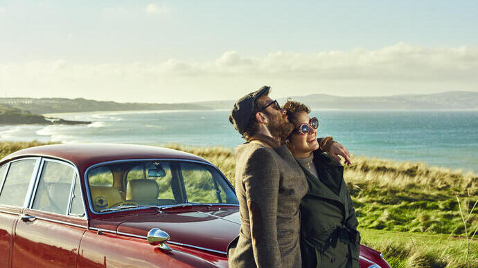 Coppia che si abbraccia e si appoggia a un'auto rossa parcheggiata in riva al mare