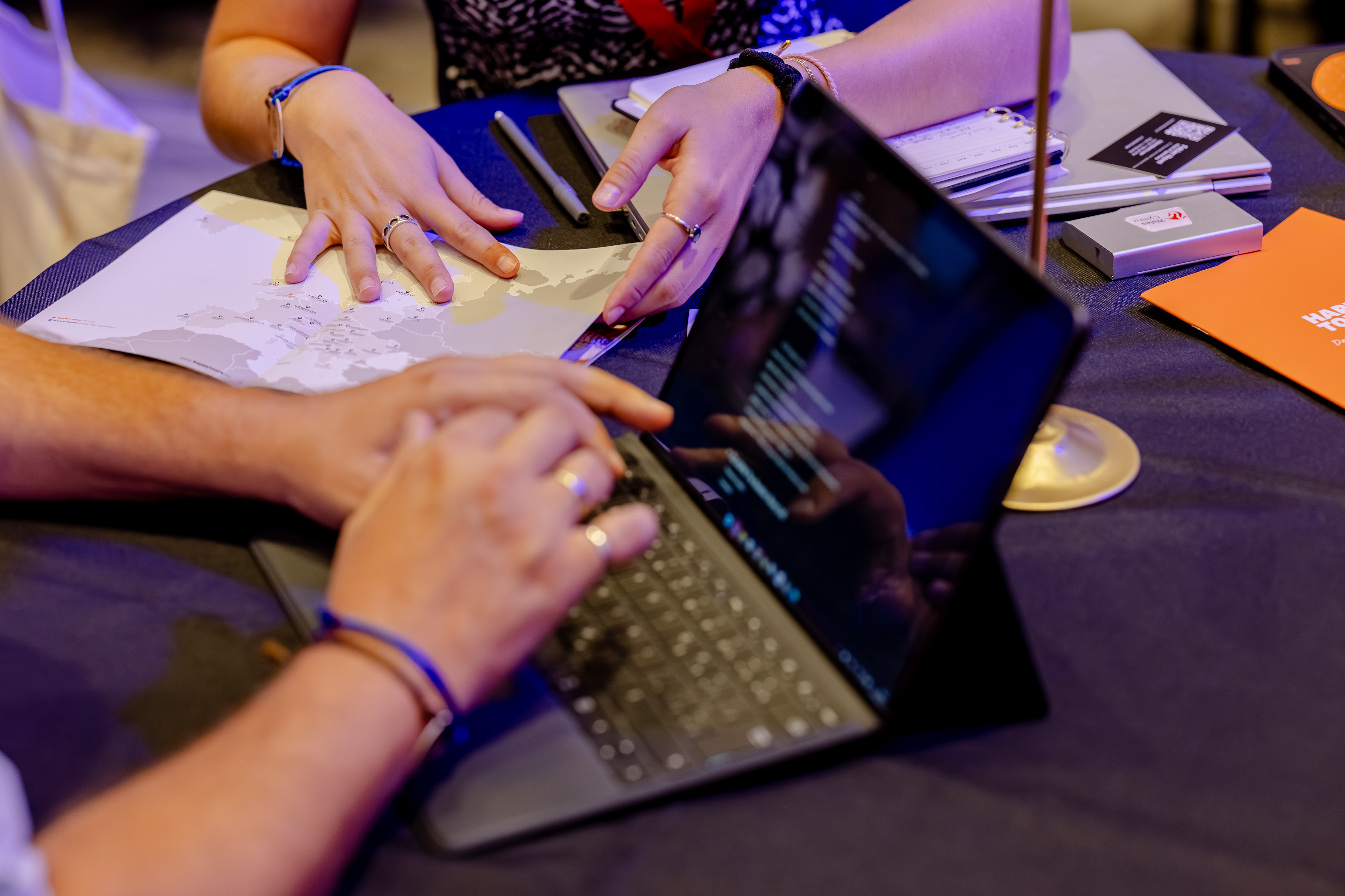 Personas trabajando en un portátil y sujetando un mapa, con bolígrafos y cuadernos sobre la mesa.