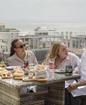 Two men and two women laugh together while sat on a balcony eating lunch with the sea beyond