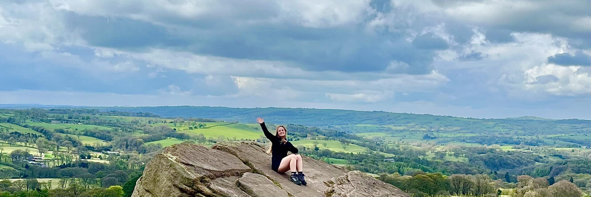 Person sitzt auf einem großen Felsen mit Blick auf eine grüne Landschaft unter bewölktem Himmel.