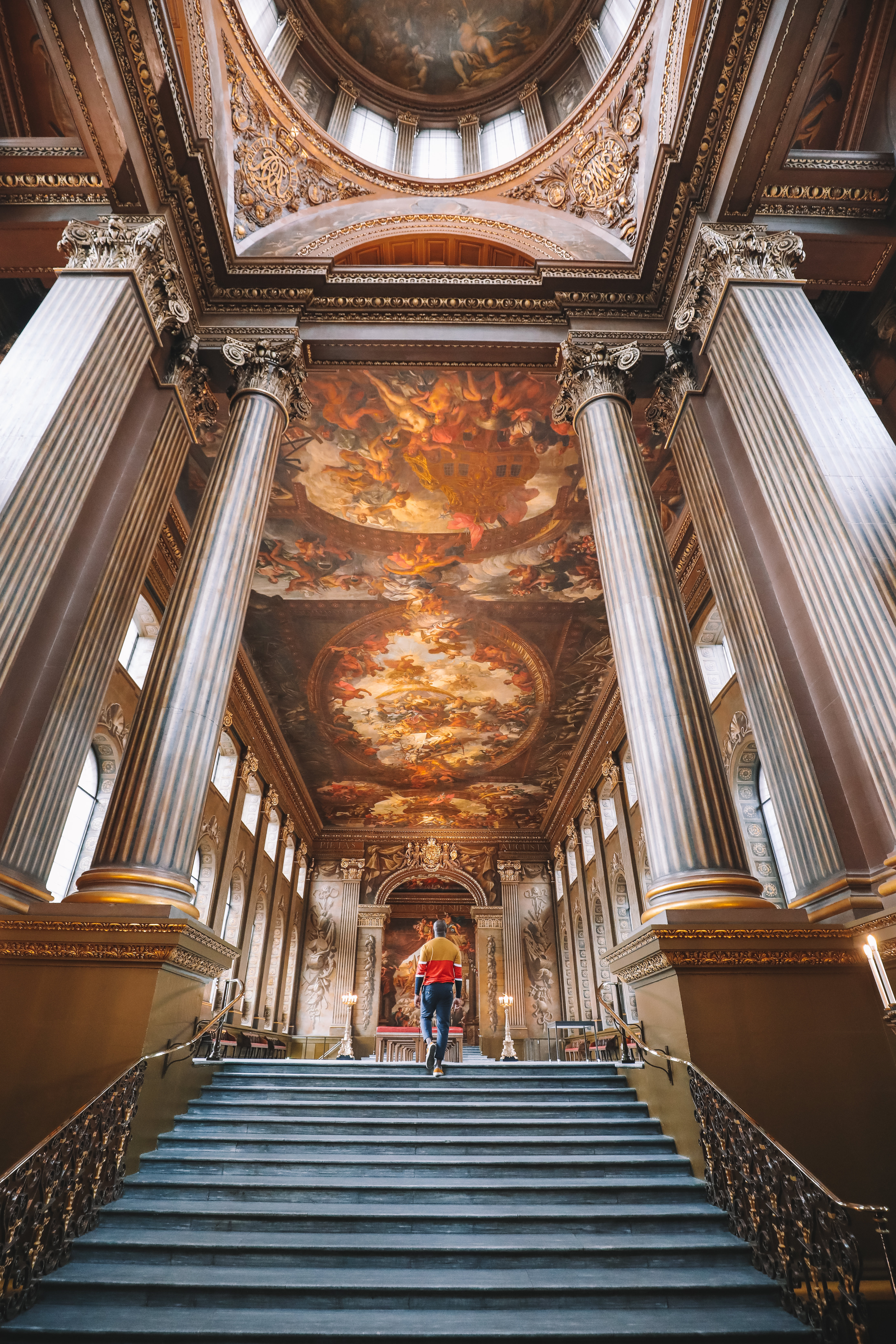 Man walking up a grand staircase towards a large hall with a painted ceiling
