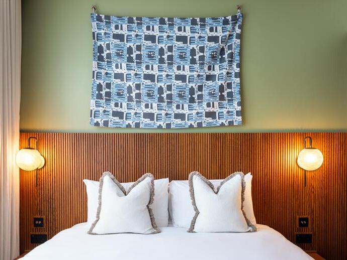 Modern hotel room with a double bed, two white pillows, wooden headboard, wall art, and two round wall lamps on either side of the bed.