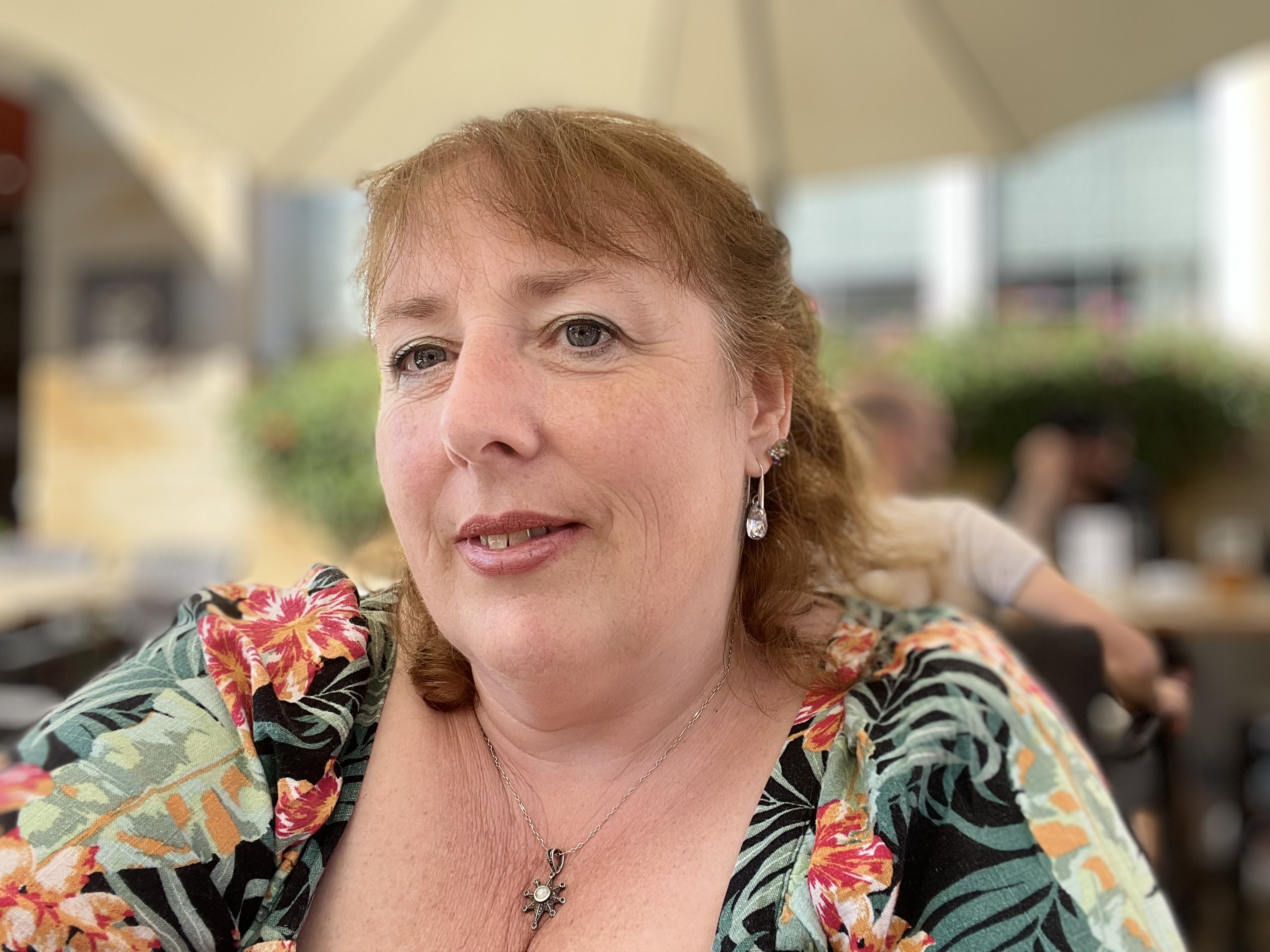 Woman with floral shirt seated outdoors under an umbrella, with blurred background of plants and people at a café or restaurant.
