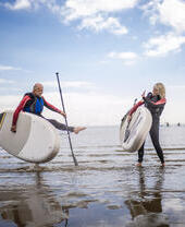 A man and woman in wet suits stand holding paddleboards on a beach