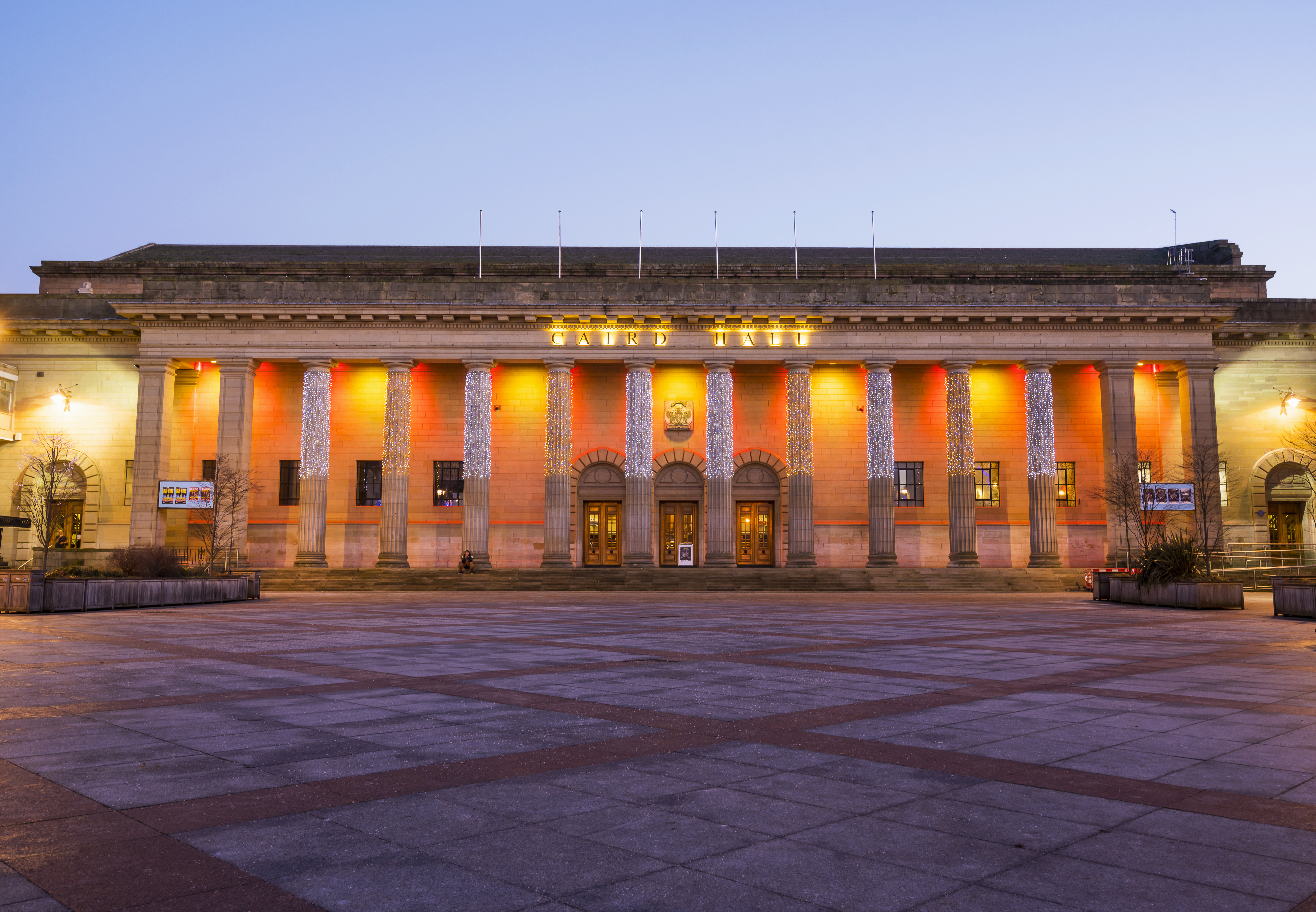 Large neoclassical building with columns, illuminated at dusk, facing an open square. Decorative lights accent the facade.