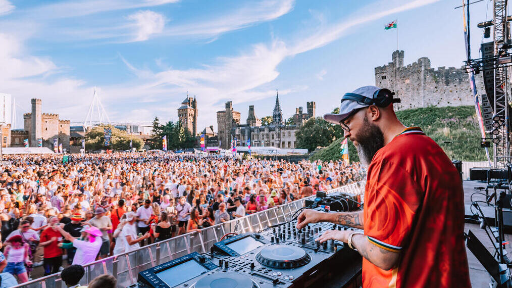 DJ spielt vor großem Publikum auf einem Musikfestival im Freien, mit einer Burg im Hintergrund und blauem Himmel.