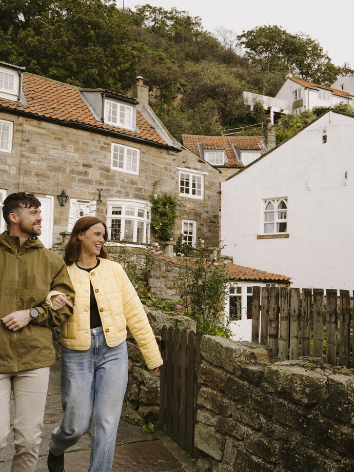 Couple walking on a cobblestone street in a quaint village with stone houses, red-tiled roofs, greenery, and hillside cottages in the background.