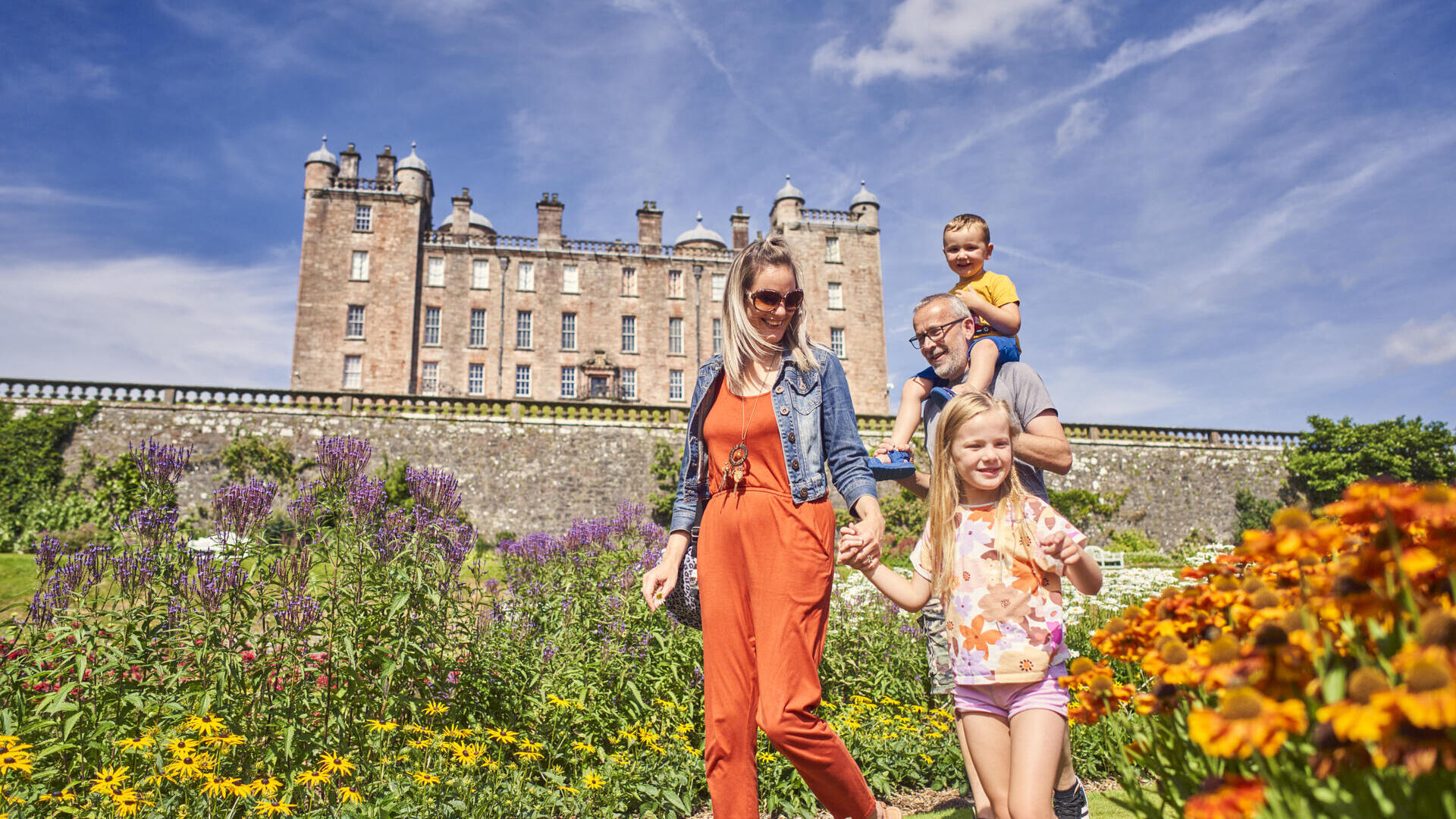 Familia caminando por un jardín con flores frente a un gran edificio histórico bajo un cielo azul claro.