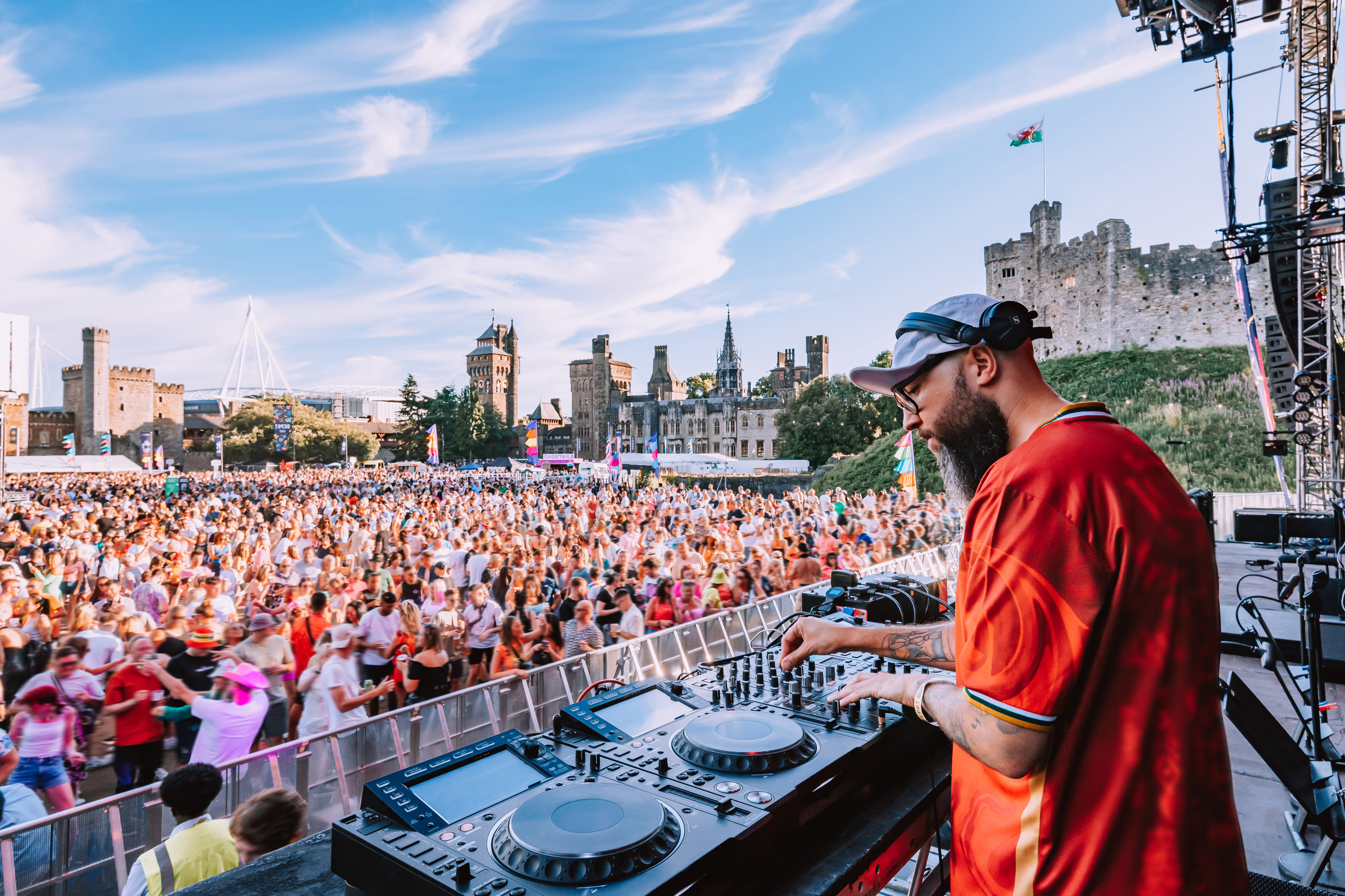 DJ spielt vor großem Publikum auf einem Musikfestival im Freien, mit einer Burg im Hintergrund und blauem Himmel.