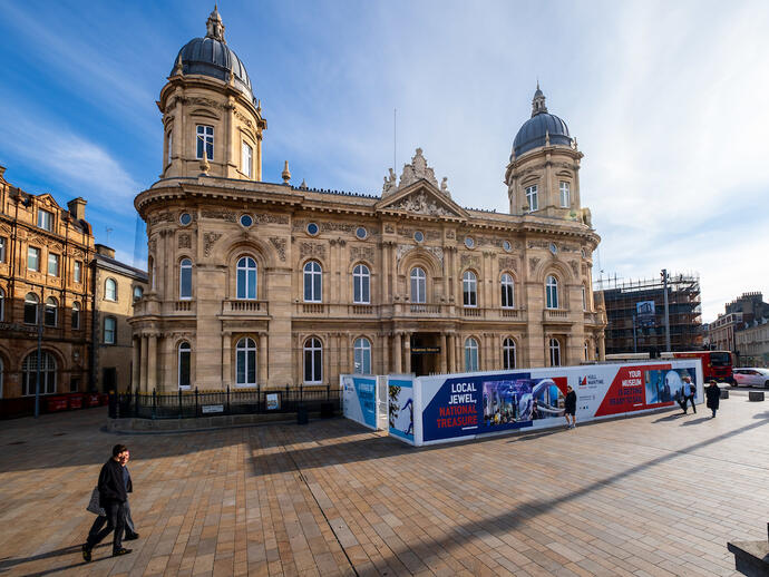 Historic domed building with ornate architecture, plaza in foreground, temporary museum construction signs, and people walking nearby on a sunny day.