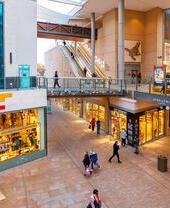 Modern indoor shopping mall with shops, escalators, colorful decor, glass railings, and people walking or shopping.