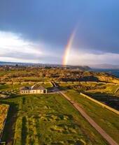 Vista aérea de una zona verde elevada sobre el mar, con un edificio pequeño y un arco iris en el cielo.