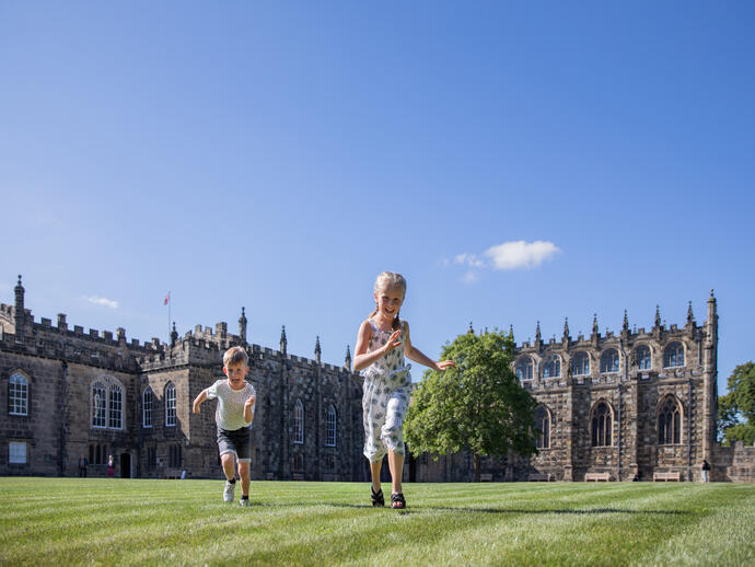 Children running and playing on grass in front of a large historic stone building under a bright blue sky.