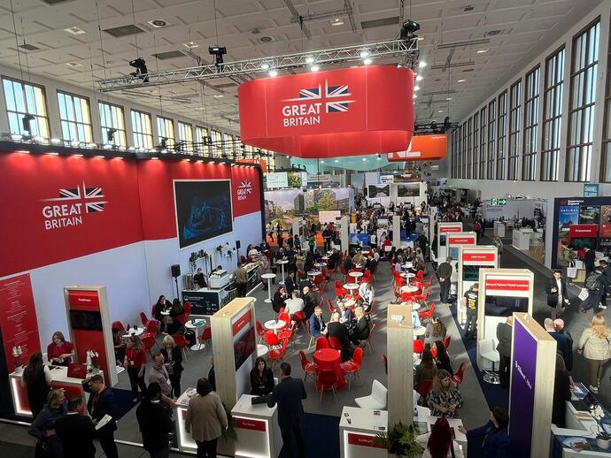 Busy trade show or exhibition hall with red and white Great Britain banners, people networking at tables, and various booths in a modern, bright venue.