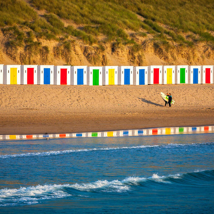 Un surfista caminando por la playa frente a coloridas casetas de playa.