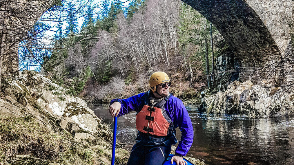 Hombre con una canoa azul de pie a orillas de un río cerca de un puente