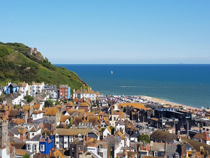 Coastal town with clustered houses, a green hill with a castle, and a beach by the blue sea under a clear sky.