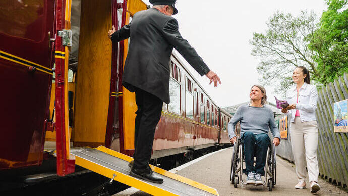 Guard stands on a ramps and leans out of heritage train carriage greeting woman and man using a wheelchair