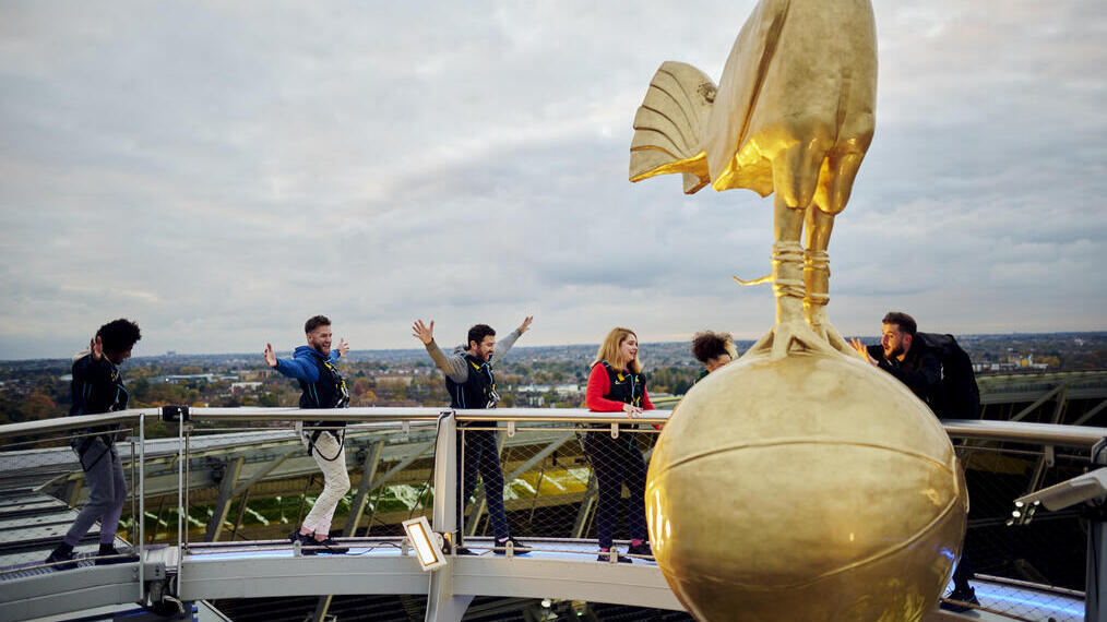Group of friends standing on a stadium roof wearing safety harnesses