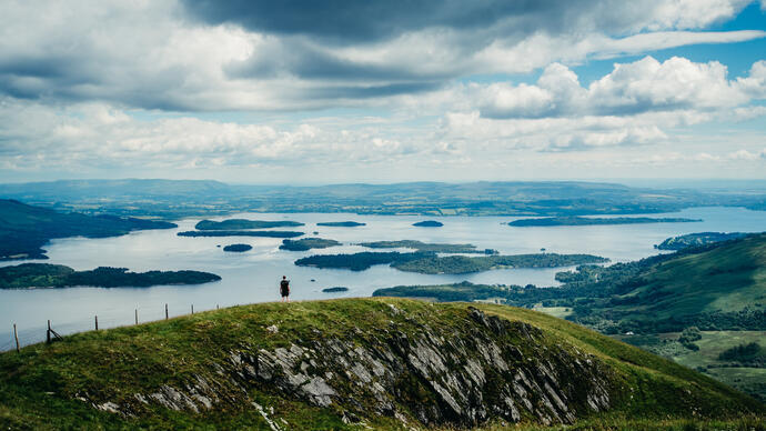 A wide panorama of a hiker standing atop a hill looking out over grassland and lakes.