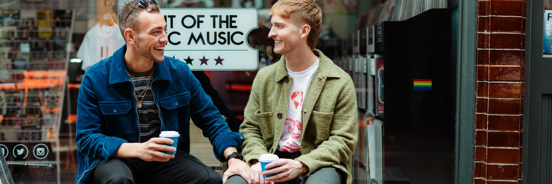Two men sit with take away drinks in front of a shop