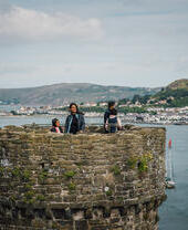 People standing atop an old stone castle tower overlooking water, sailboats, and a coastal town with hills in the background under a cloudy sky.