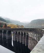 Two people walking on an arched bridge spanning a lake, with a valley in the background