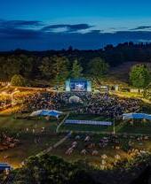A large outdoor concert with a large screen with crowds and many tents and fairground rides.