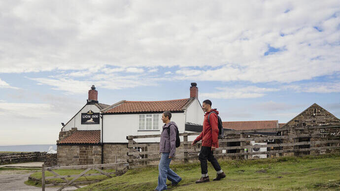 Two people walk on grass past an old inn with red-tiled roof and stone walls under a partly cloudy sky. Wooden fencing and rural scenery are visible.