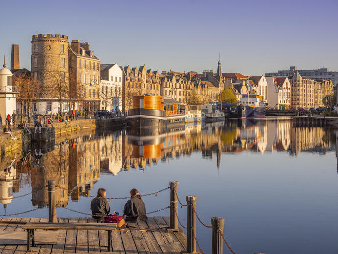 Two young women sat on the jetty of the waterfront in the evening with boats and buildings by the harbour.