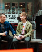 Two men sit with take away drinks in front of a shop