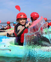 Children in helmets and life jackets kayaking on a lake under a clear blue sky, water splashing in foreground.