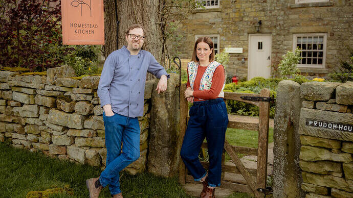 Dos personas delante de una casa rural. Un cartel dice: «Homestead Kitchen»