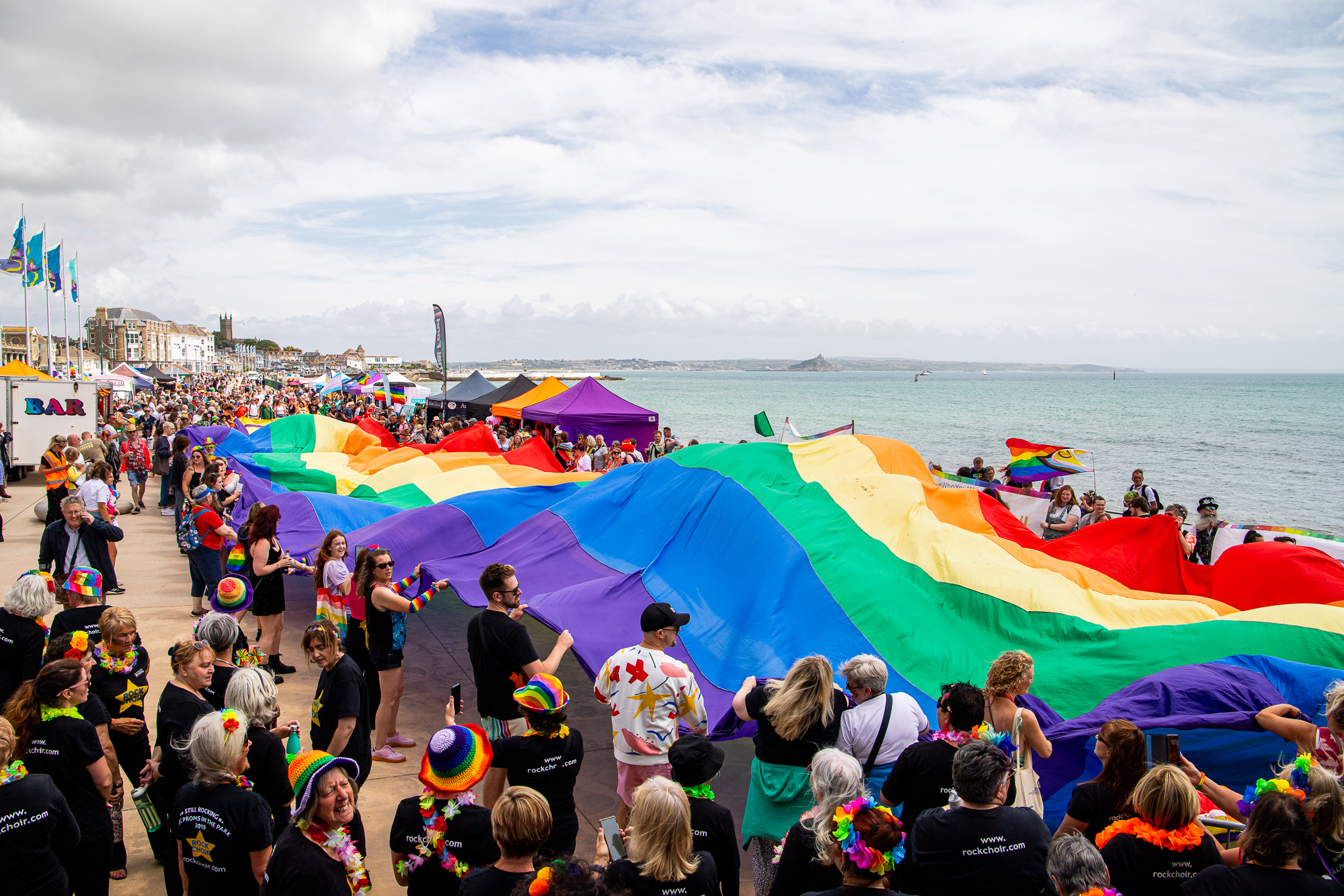 Eine Menschenmenge hält eine große Pride-Flagge an der Küste von Cornwall hoch