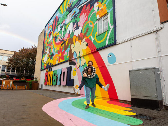 Two people playfully smiling and posing in front of a colourful mural.