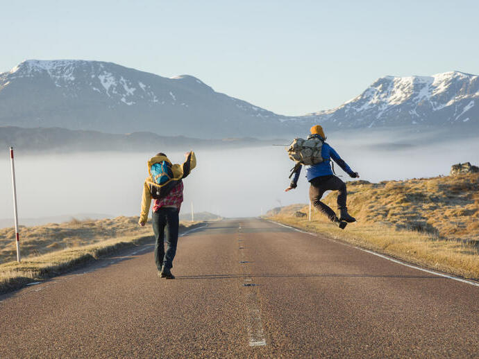 Two men walking and jumping on a road surrounded by mountains