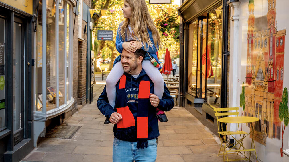 Mann mit einem Kind auf den Schultern spaziert durch eine Fußgängerzone mit Geschäften auf beiden Seiten