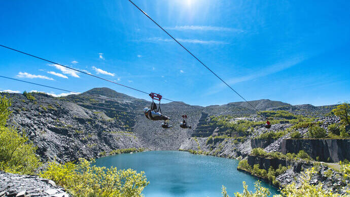 People ziplining over a blue lake in a rocky quarry landscape under a clear sunny sky, surrounded by hills and scattered greenery.