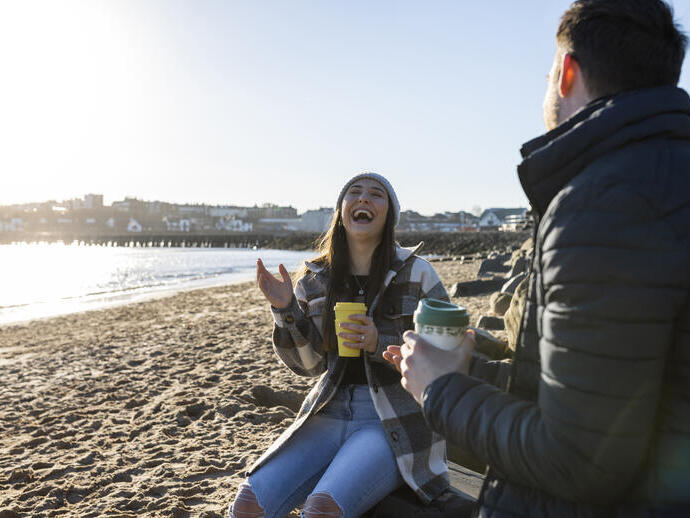 Two people sit on a sandy beach at sunset, laughing and holding takeaway coffee cups, with seaside buildings in the background.