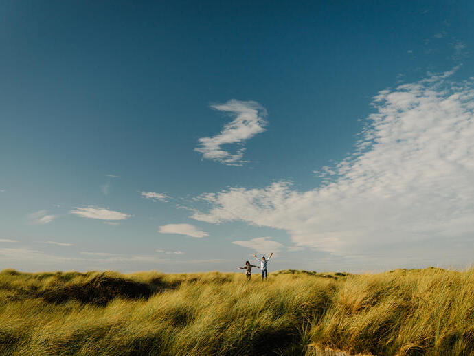 Twee mensen lopen door hoog gras onder een blauwe lucht met enkele wolken.